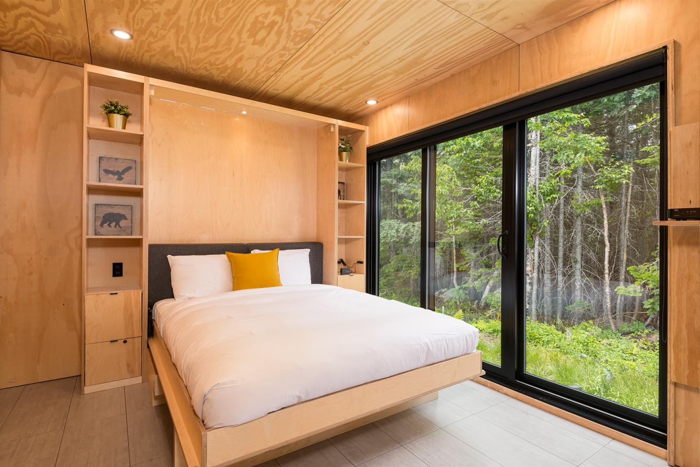 Chambre moderne en bois clair avec grande baie vitrée donnant sur une forêt.