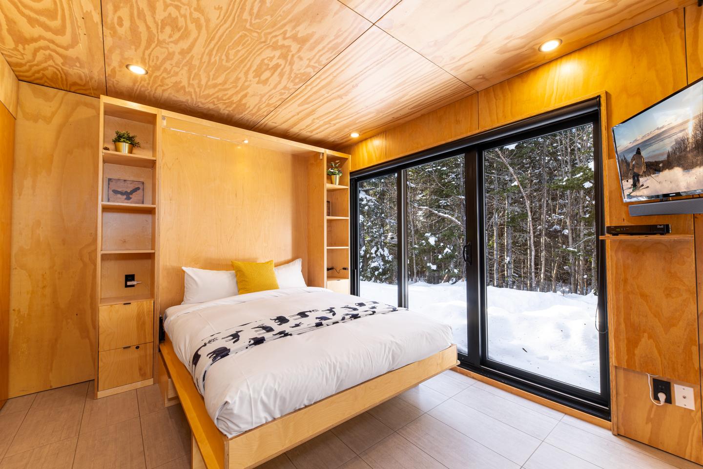 Chambre en bois clair, lit double, grandes fenêtres donnant sur la neige et forêt.