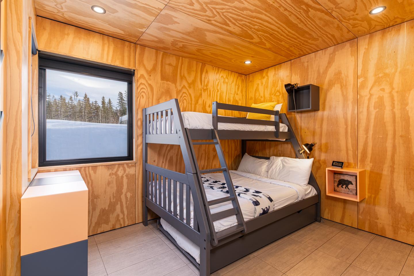 Chambre en bois avec lits superposés, fenêtre, et étagère murale.