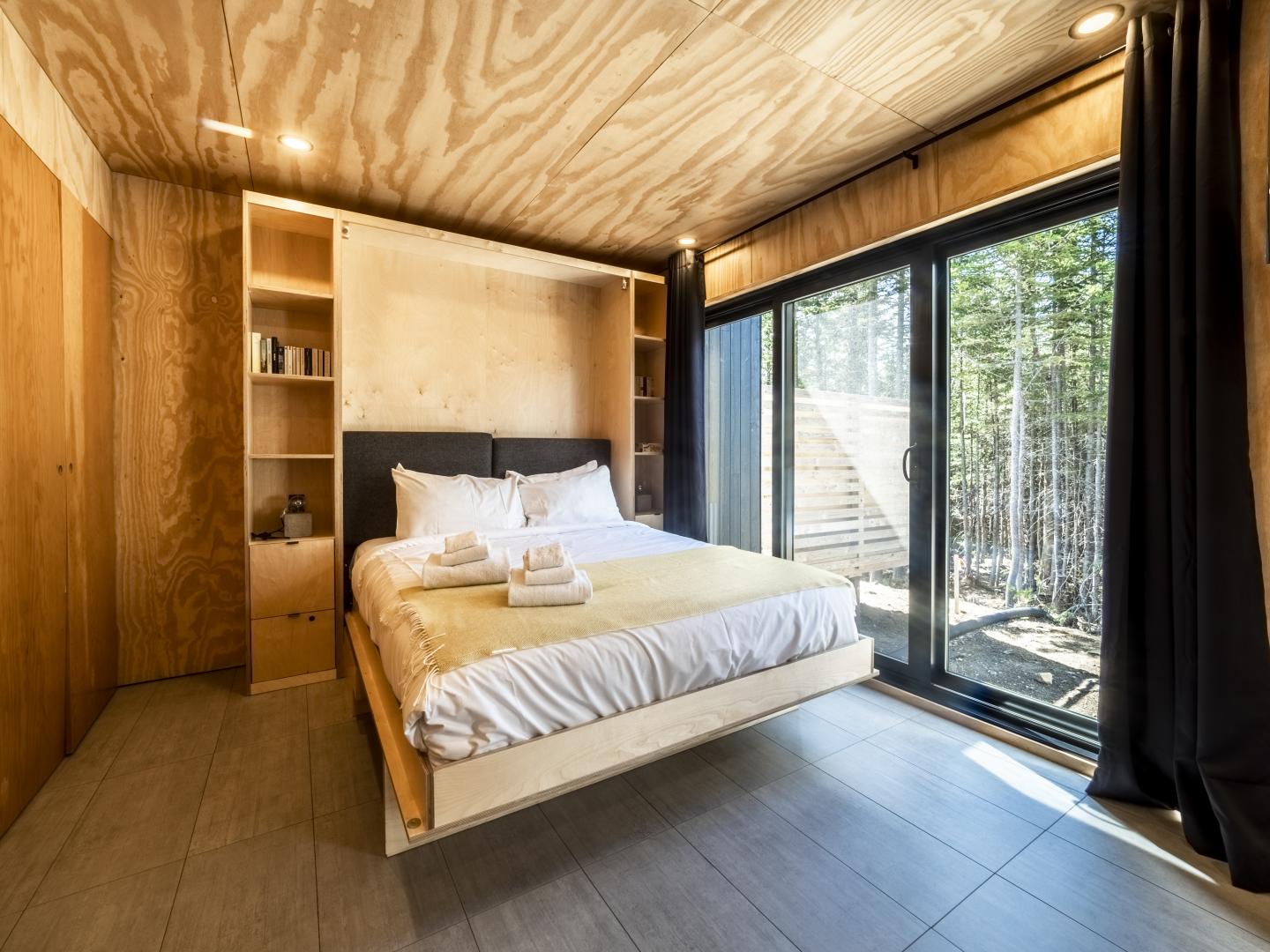 Chambre moderne avec lit, grandes fenêtres, bois clair et vue sur la forêt.