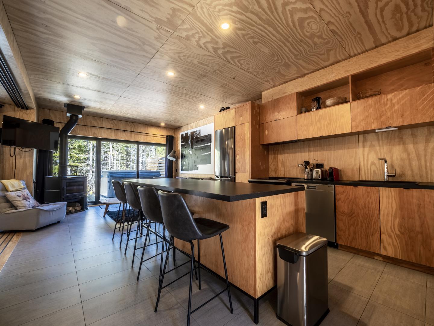 Cuisine moderne en bois, avec îlot central, tabourets noirs et grandes fenêtres lumineuses.