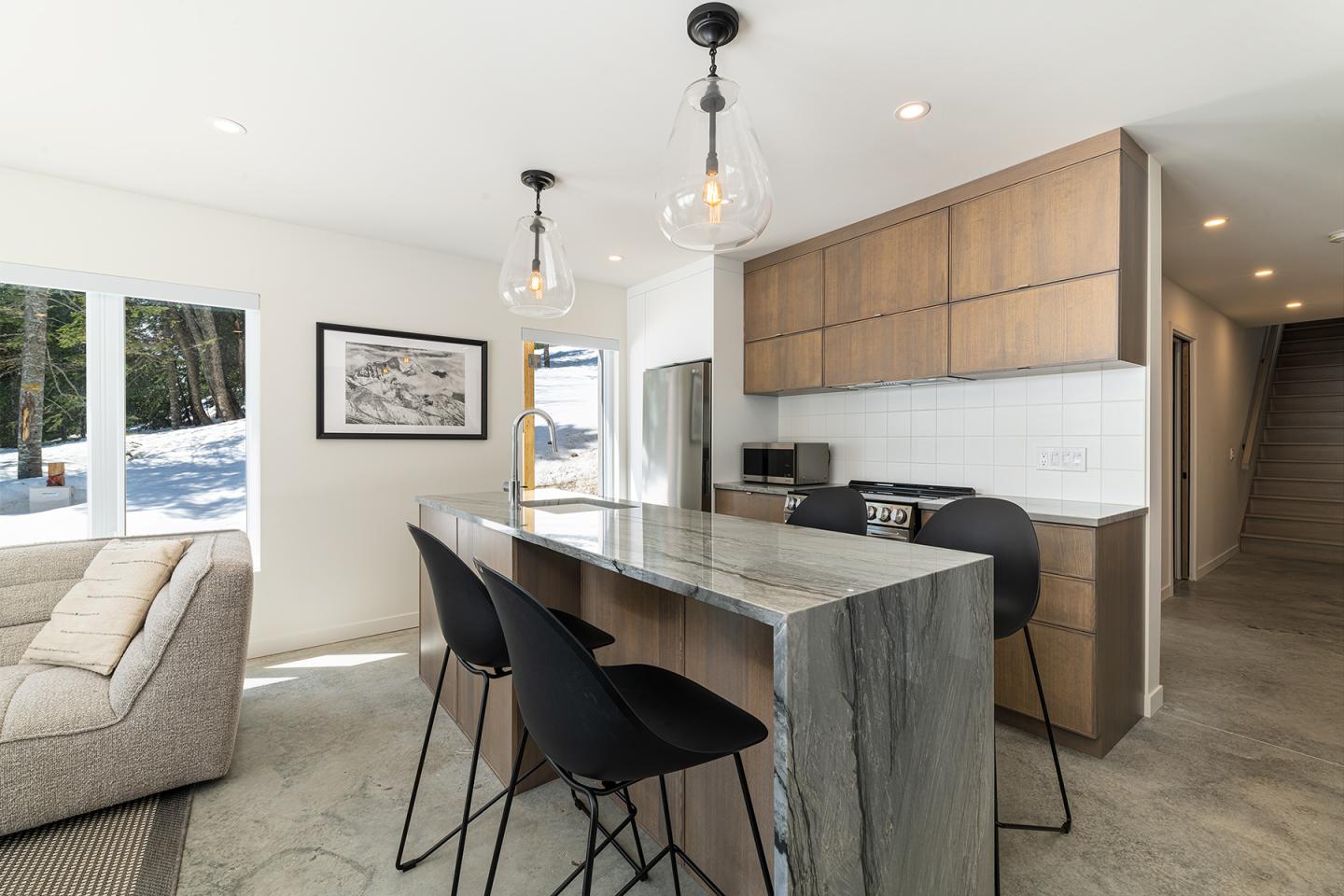Cuisine moderne avec îlot en marbre, chaises noires, et luminaires suspendus.