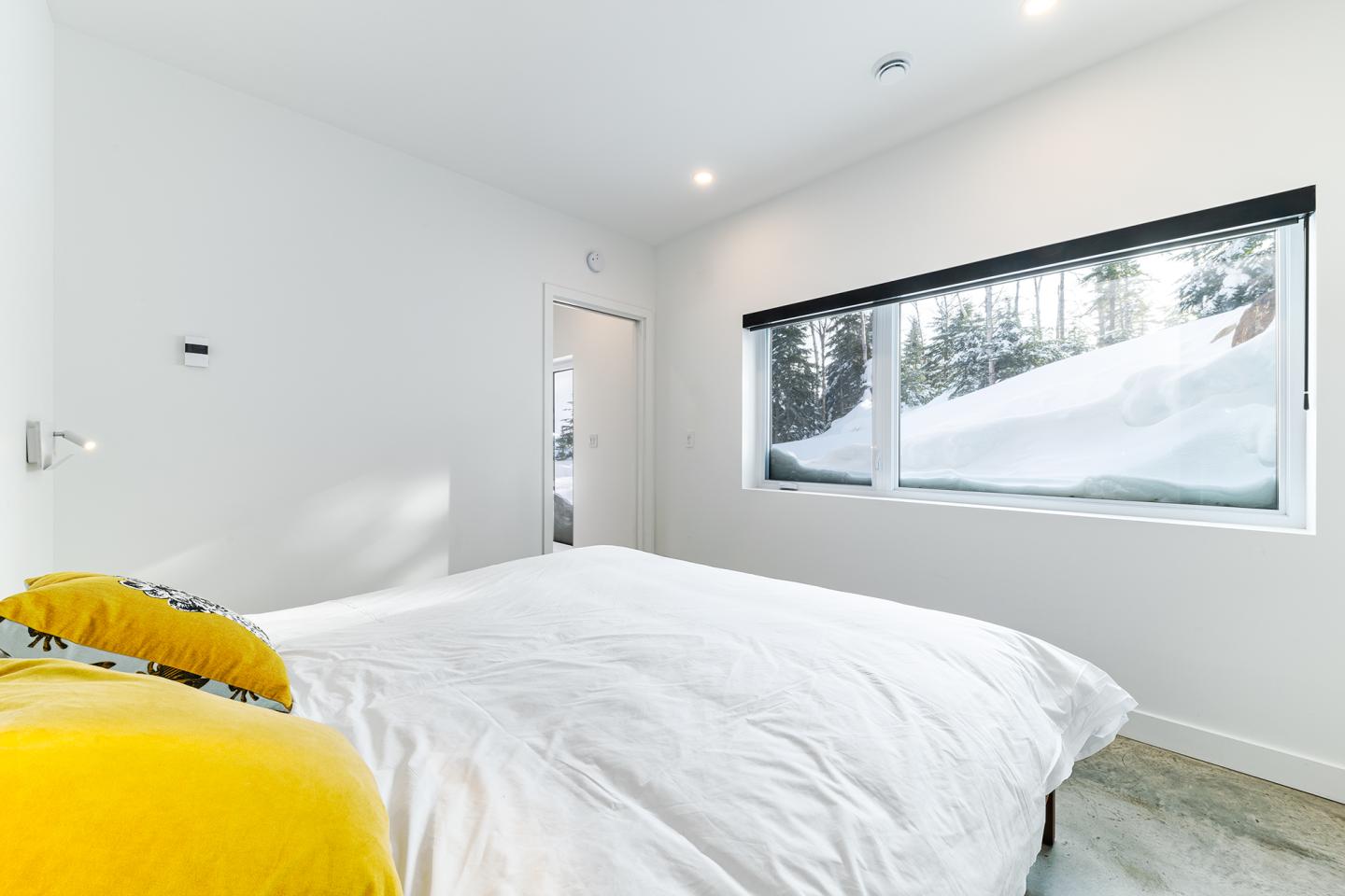 Chambre minimaliste blanche, oreiller jaune, grande fenêtre vue sur neige.