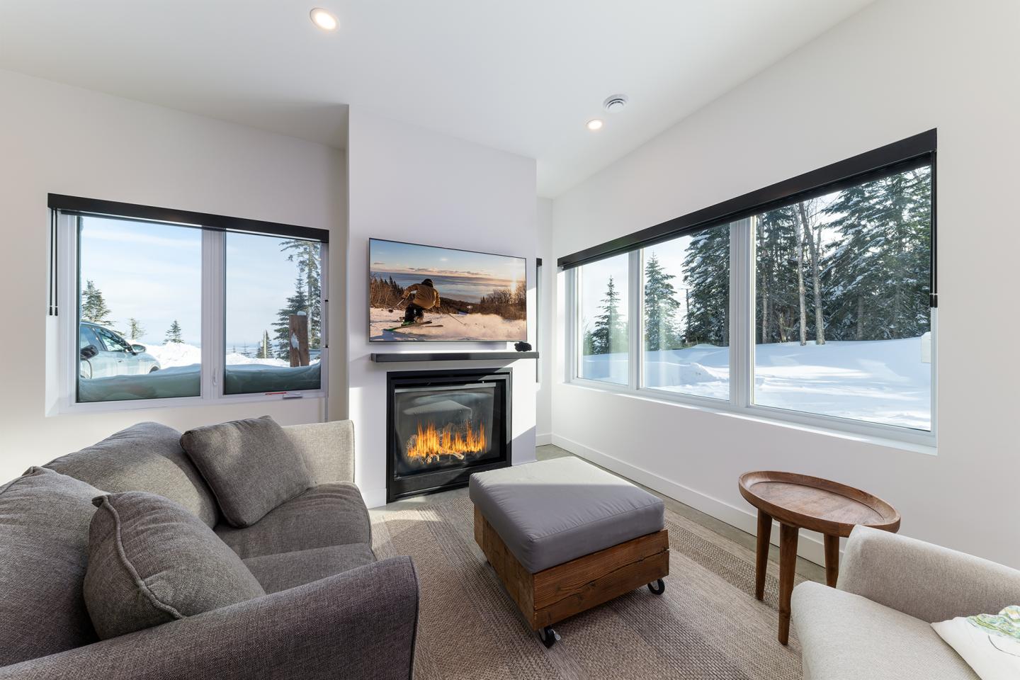 Salon moderne avec cheminée, canapé gris, vue sur la neige par les fenêtres.