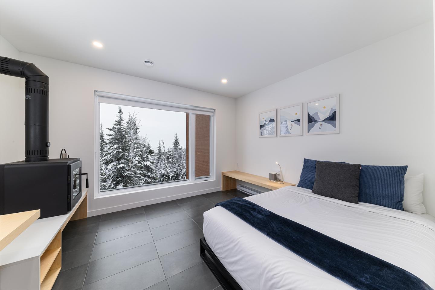 Chambre moderne avec grande fenêtre, vue sur forêt enneigée, lit et poêle à bois.