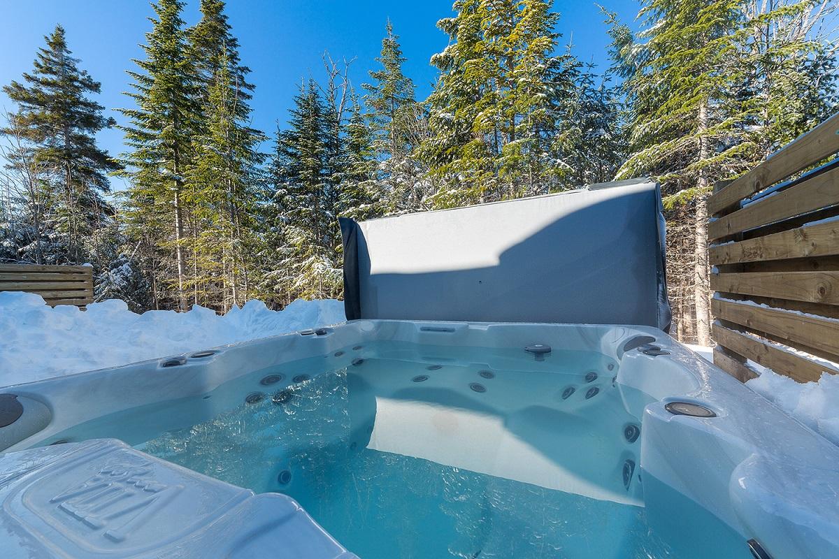 Jacuzzi extérieur entouré de neige et de sapins sous un ciel bleu.