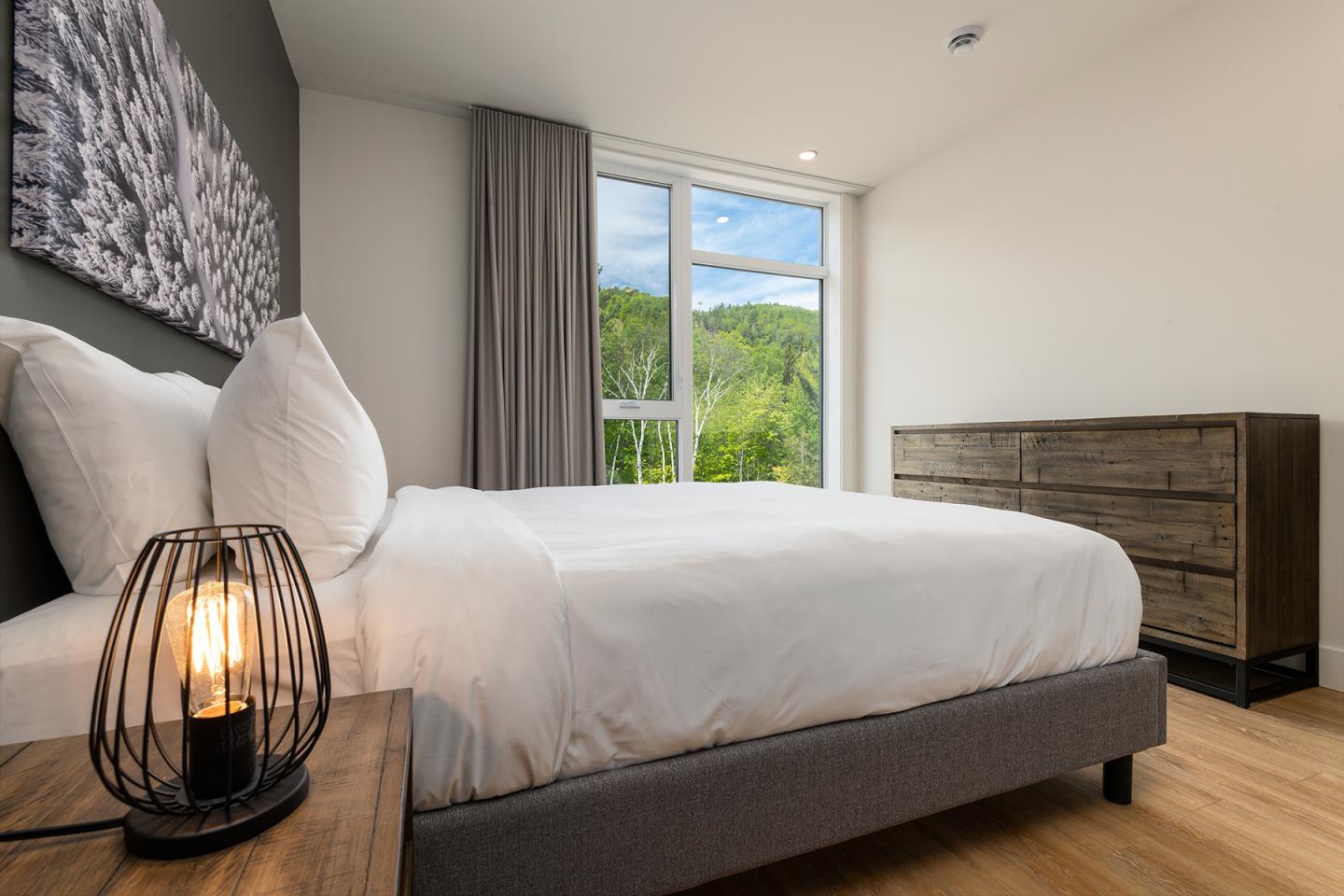 Chambre moderne avec lit double, lampe de chevet et fenêtre donnant sur des arbres.