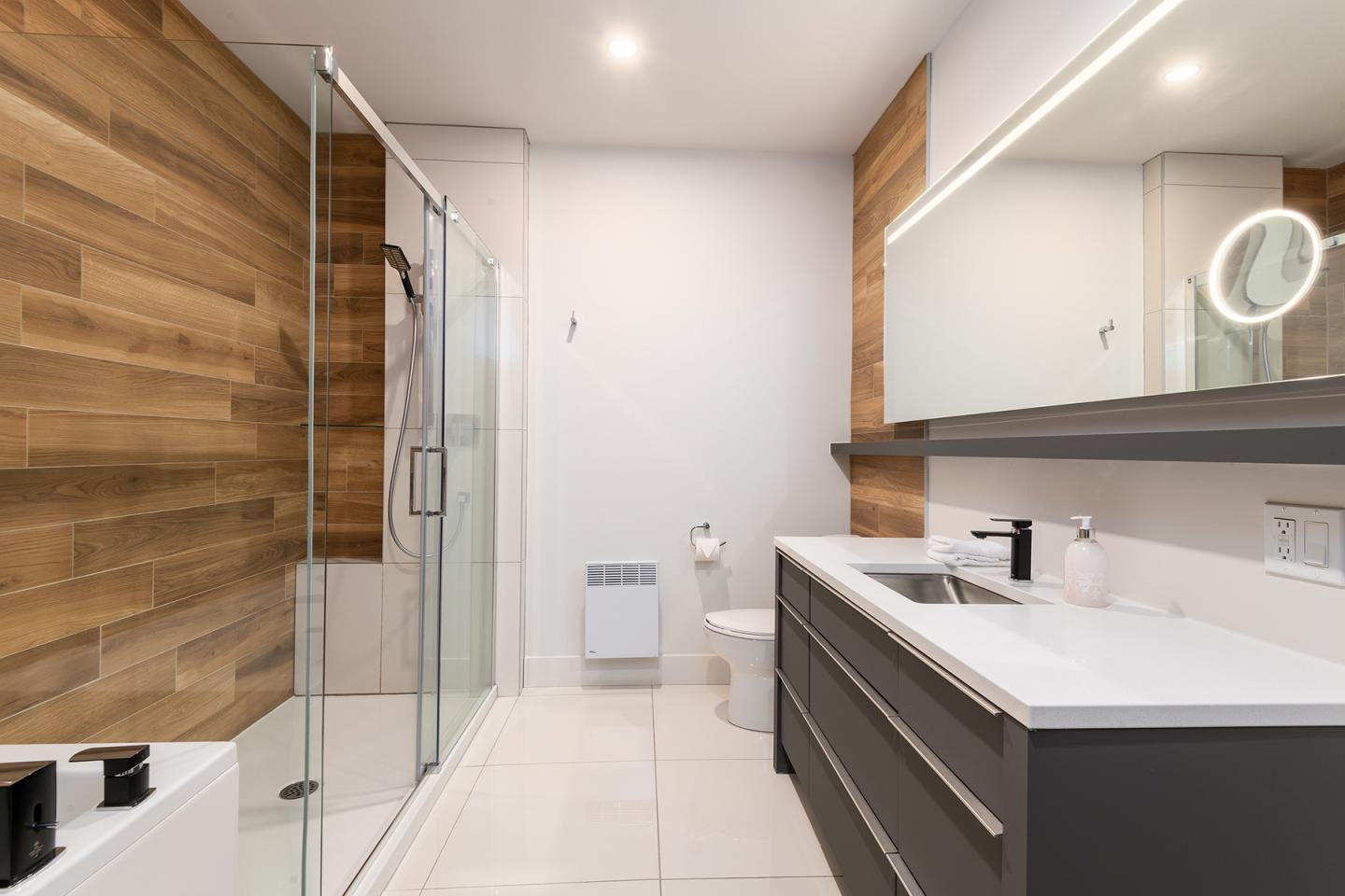 Salle de bain moderne avec douche vitrée, murs en bois, et grand miroir lumineux.