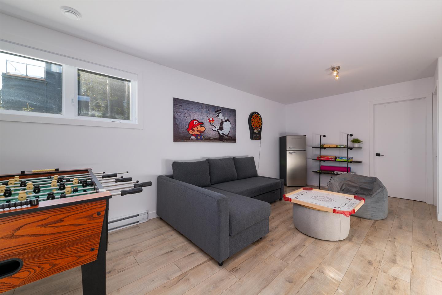 Salon moderne avec canapé gris, baby-foot et étagère colorée. Lumineux et épuré.