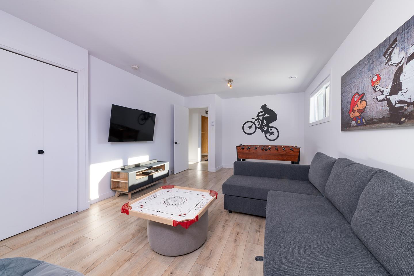 Salle de jeux moderne avec canapé gris, télé, baby-foot et poster de cycliste.