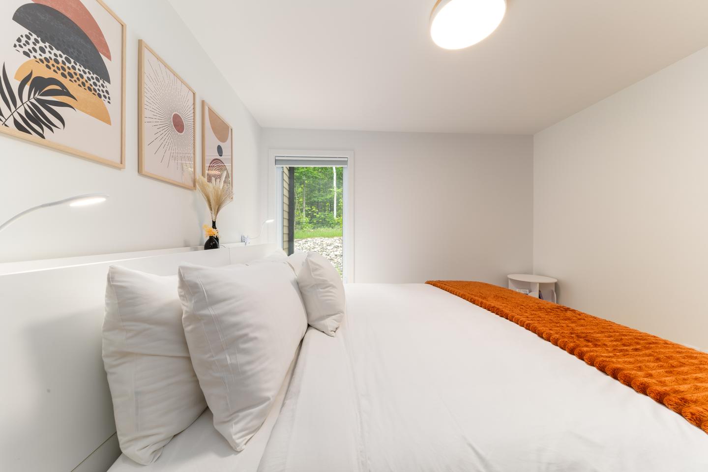 Chambre moderne avec lit blanc, plaid orange et trois tableaux au mur.