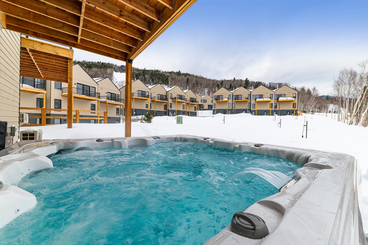Jacuzzi extérieur entouré de neige, vue sur des chalets et montagnes.