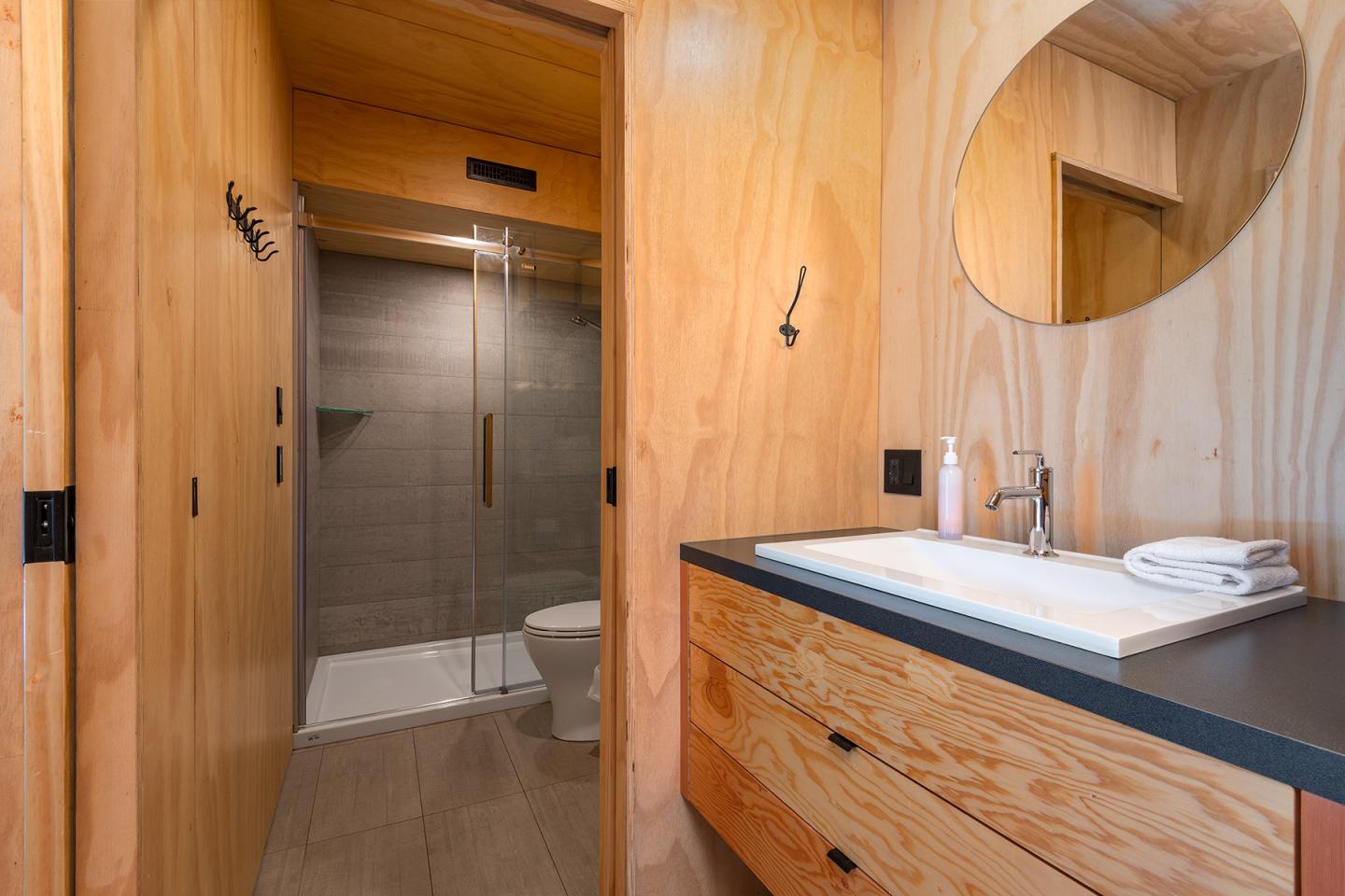 Salle de bain en bois clair avec douche, lavabo et miroir rond.