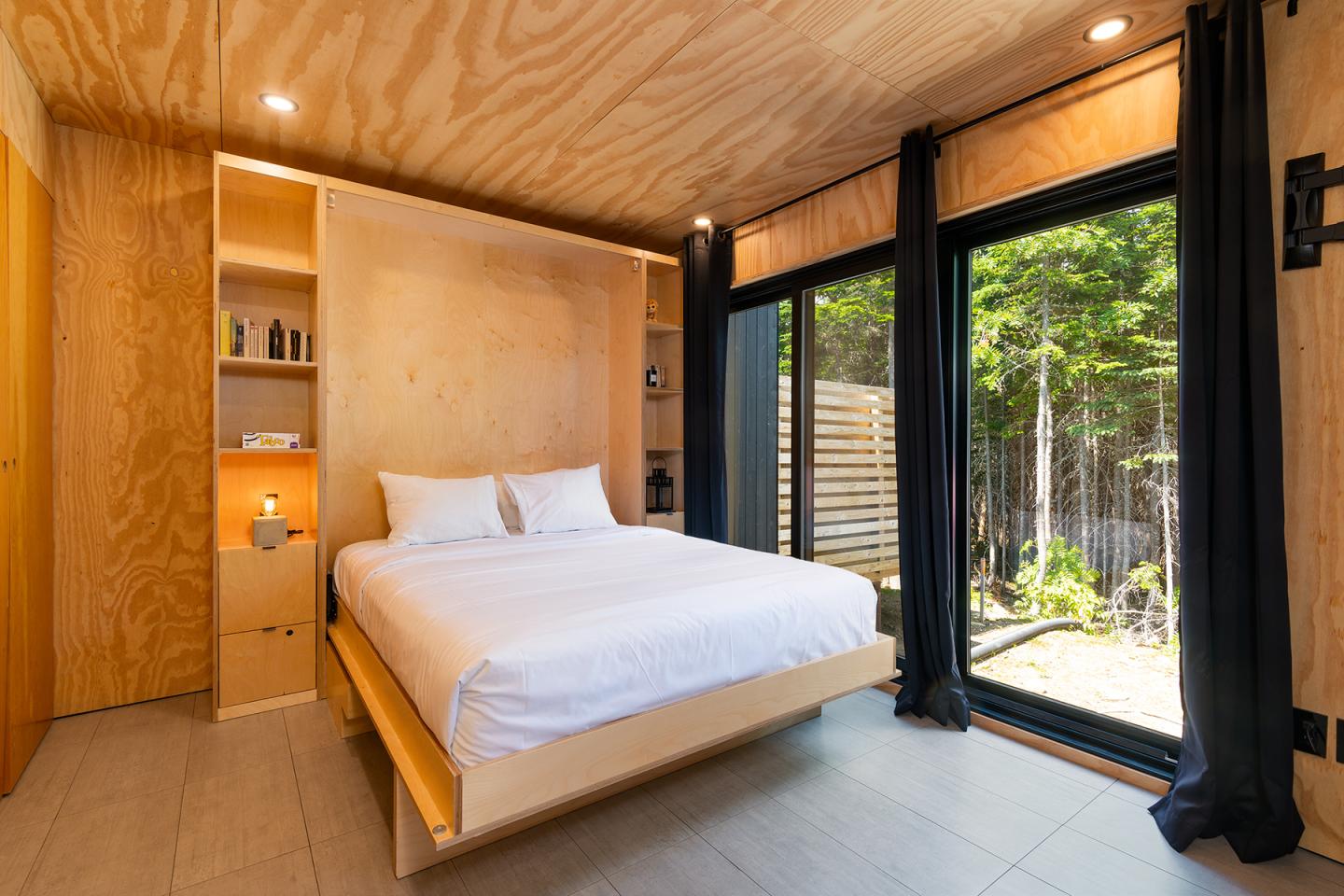 Chambre en bois clair, lit double, grande fenêtre sur forêt.