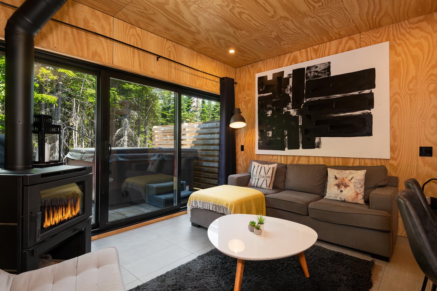 Salon moderne avec poêle, canapé gris, et table basse blanche, vue sur terrasse boisée.