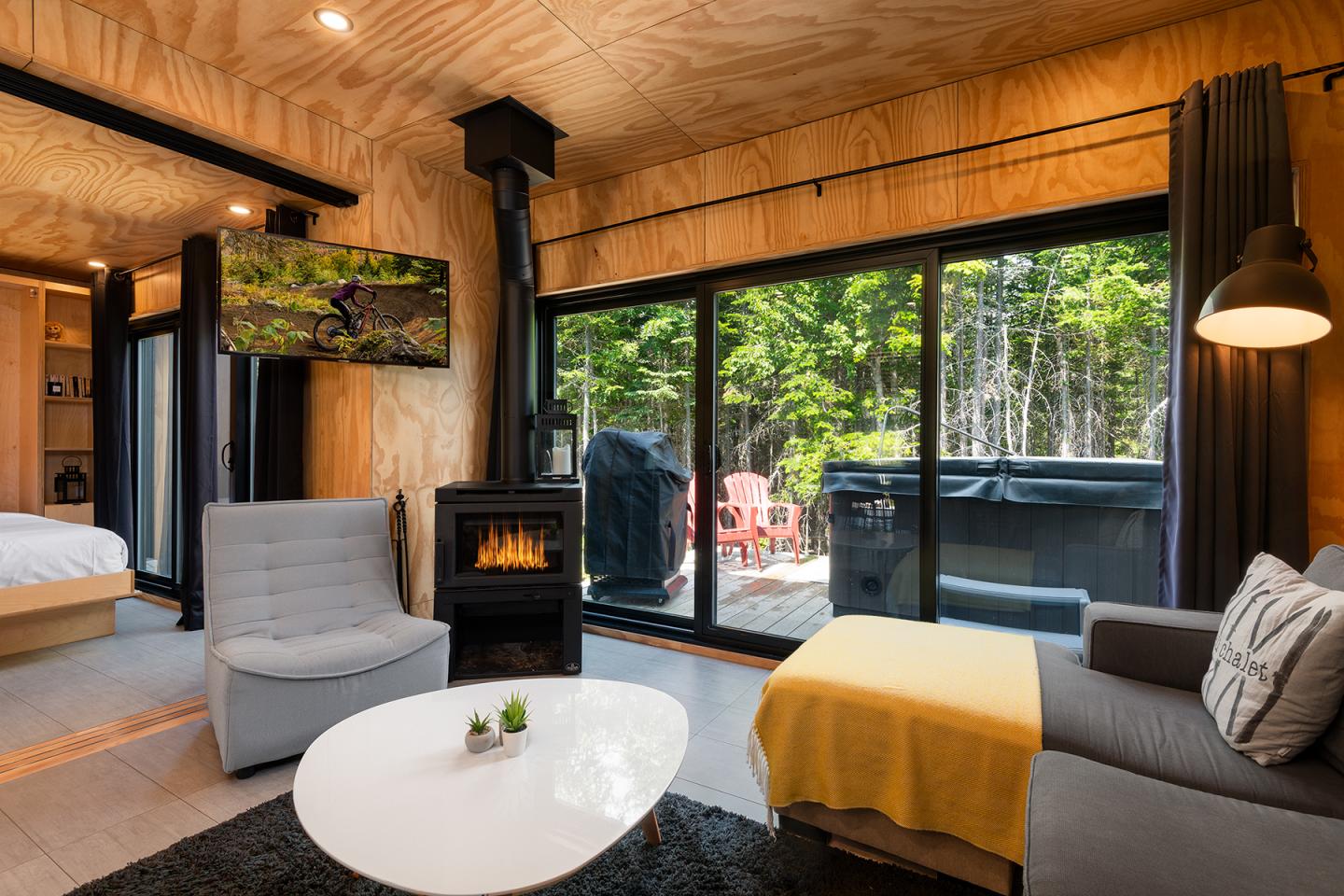 Salon moderne avec cheminée, canapé gris et vue forêt par baie vitrée.