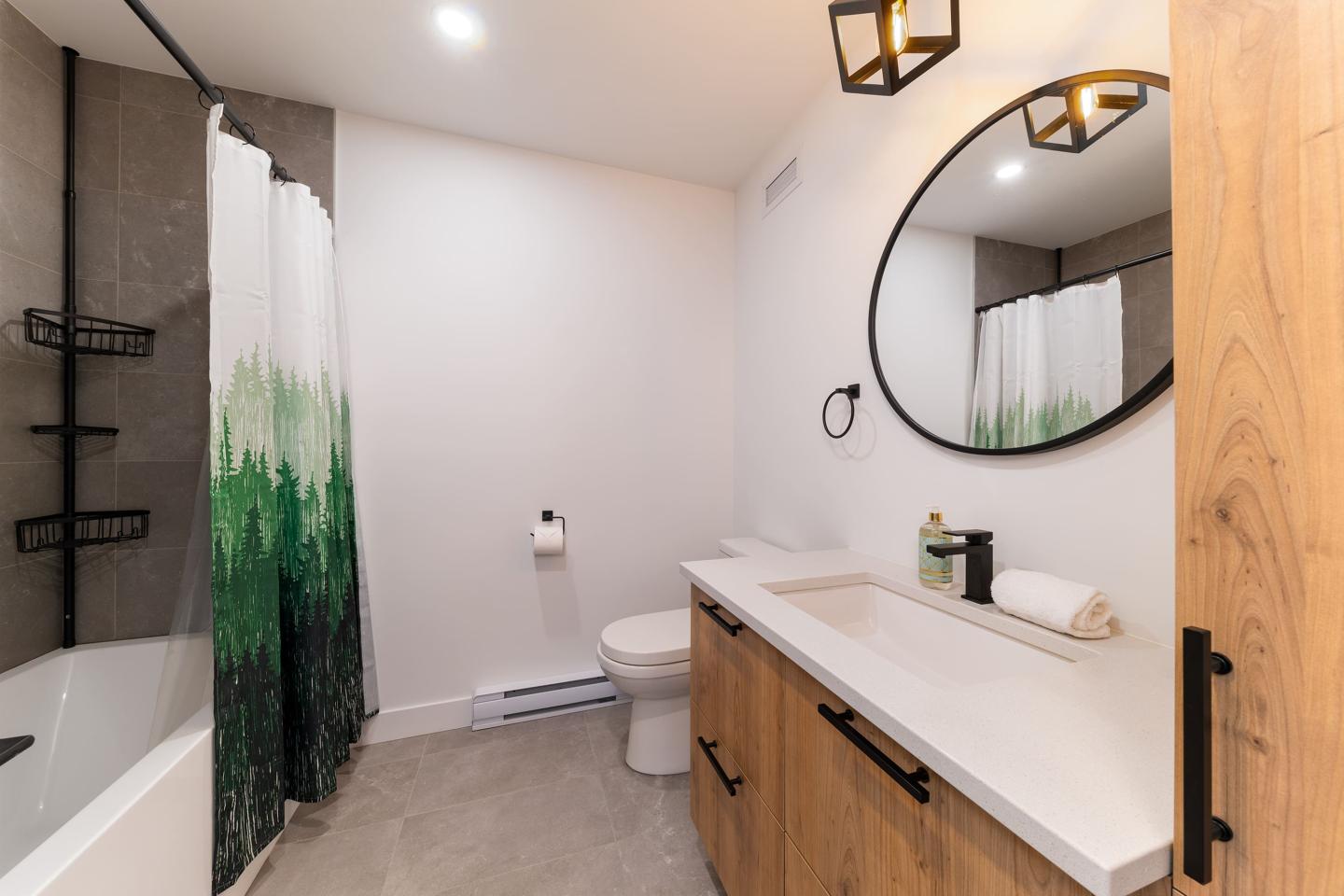 Salle de bain moderne avec baignoire, rideau vert, miroir rond et meubles en bois clair.