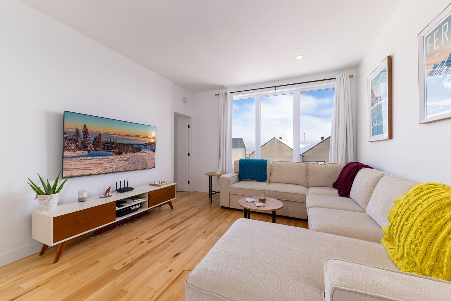 Salon moderne avec canapé beige, télé, grande fenêtre et parquet clair.