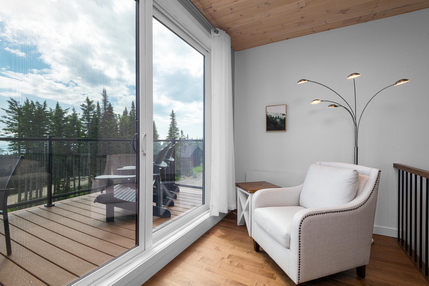 Salon lumineux avec fauteuil blanc, vue sur terrasse en bois et forêt par grande baie vitrée.