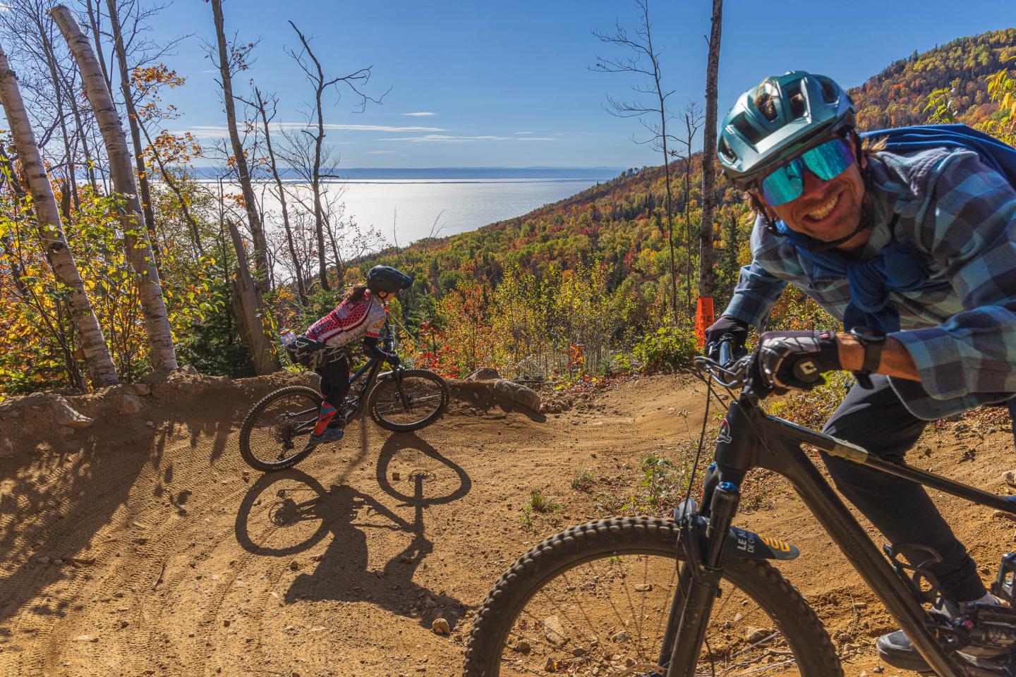 Cyclistes souriants sur un sentier forestier, lac en arrière-plan, journée ensoleillée.