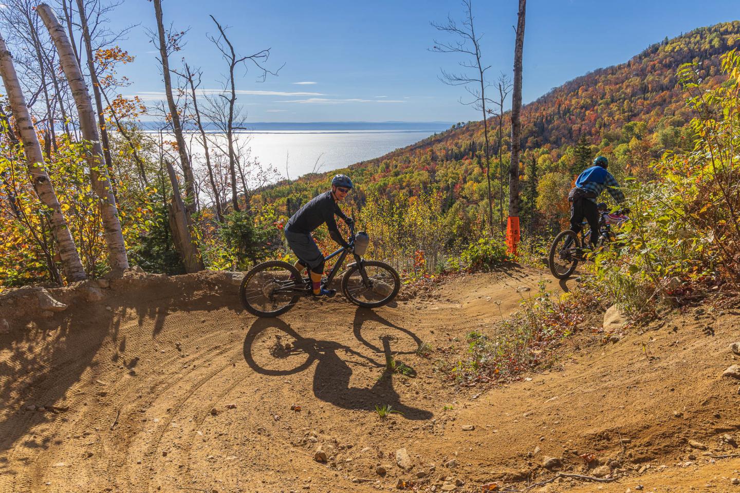 Deux cyclistes descendant un sentier de forêt en automne, vue sur lac.