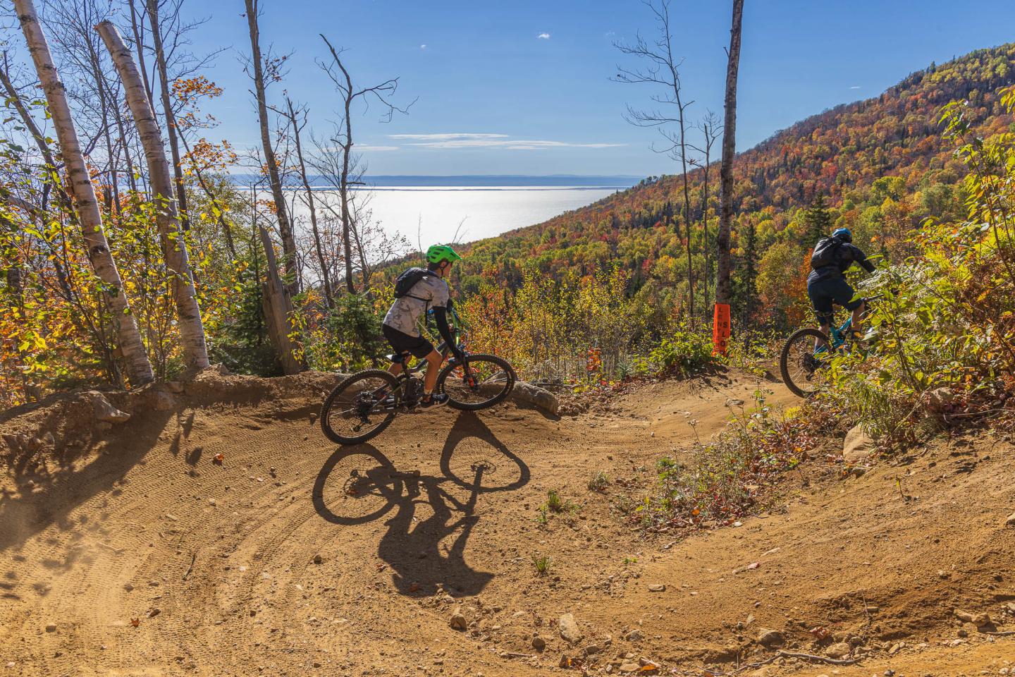Deux cyclistes descendent un sentier de montagne en automne, vue sur un lac en arrière-plan.