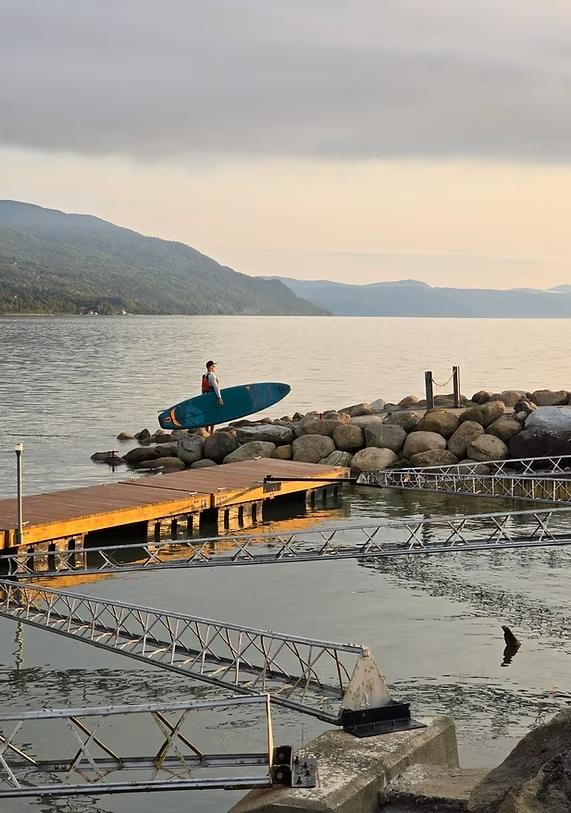 Surfeur avec planche, au bord d'un lac calme, sur des rochers sous un ciel nuageux.