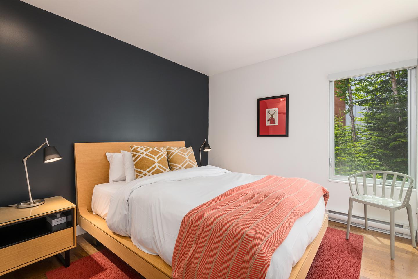 Chambre moderne avec lit double, mur noir, lampe de chevet et fenêtre donnant sur des arbres.