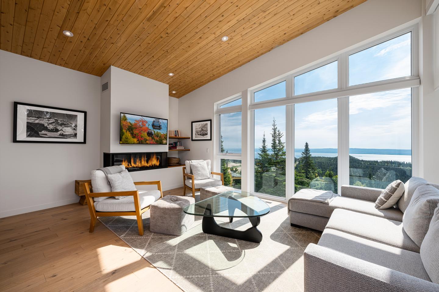 Salon moderne avec grandes fenêtres, vue sur mer, canapé gris et cheminée allumée.