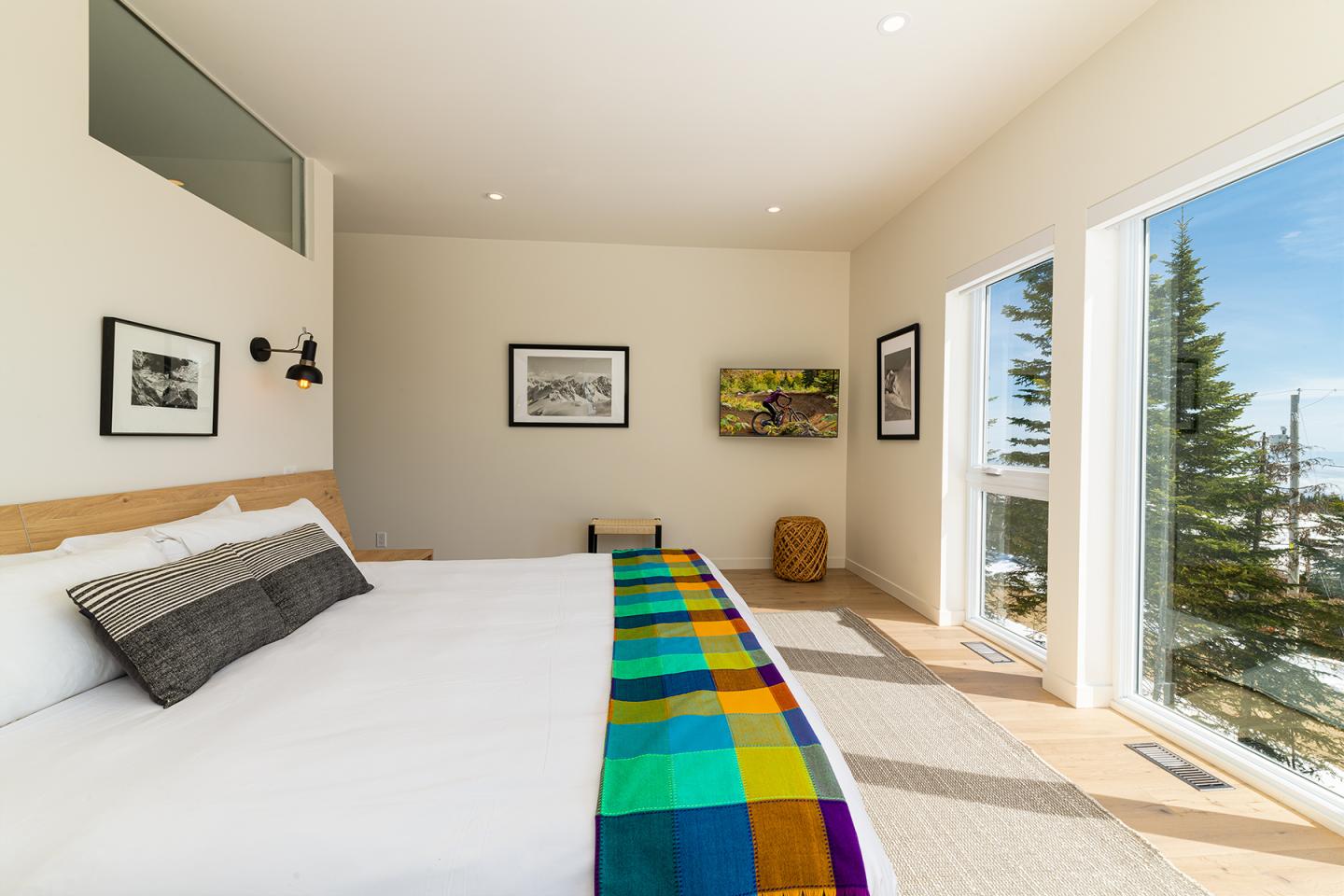 Chambre lumineuse avec lit, plaid coloré et grande fenêtre donnant sur des arbres.