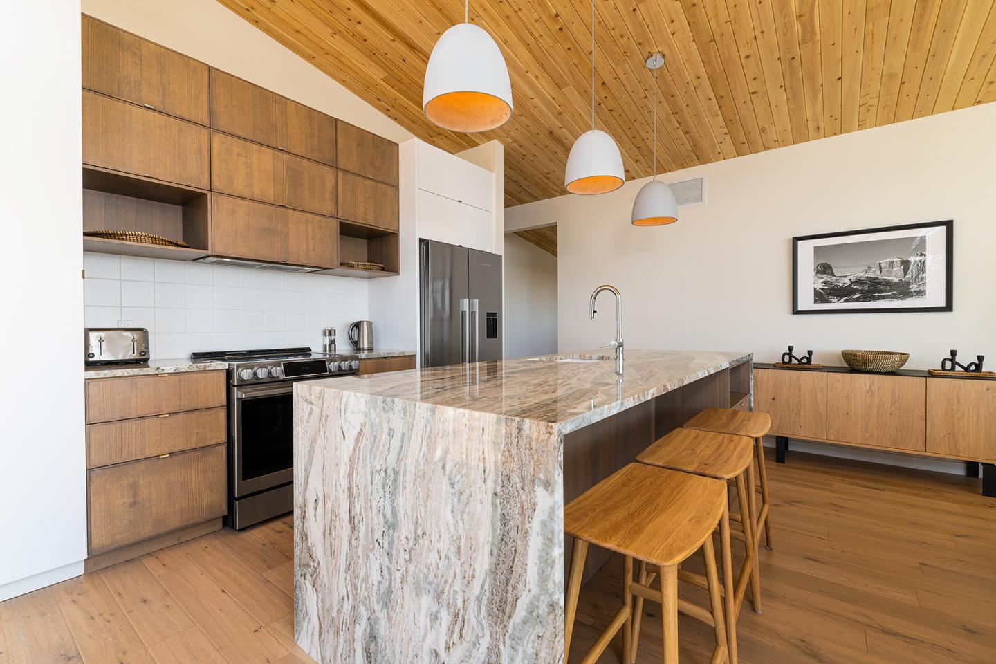 Cuisine moderne avec îlot en marbre, chaises en bois et plafonds lambrissés.