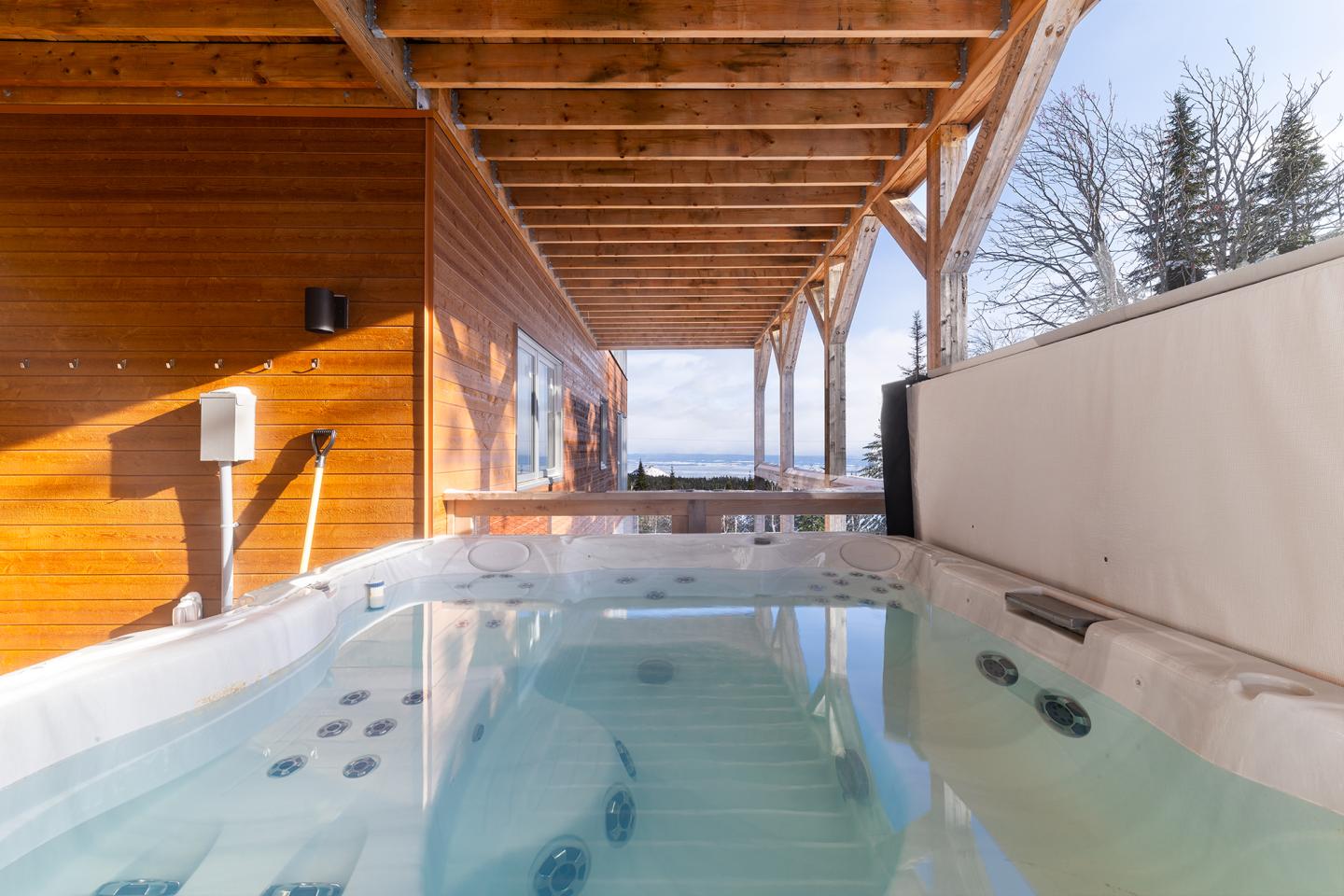 Jacuzzi extérieur sous une terrasse en bois, ciel bleu, arbres au loin.