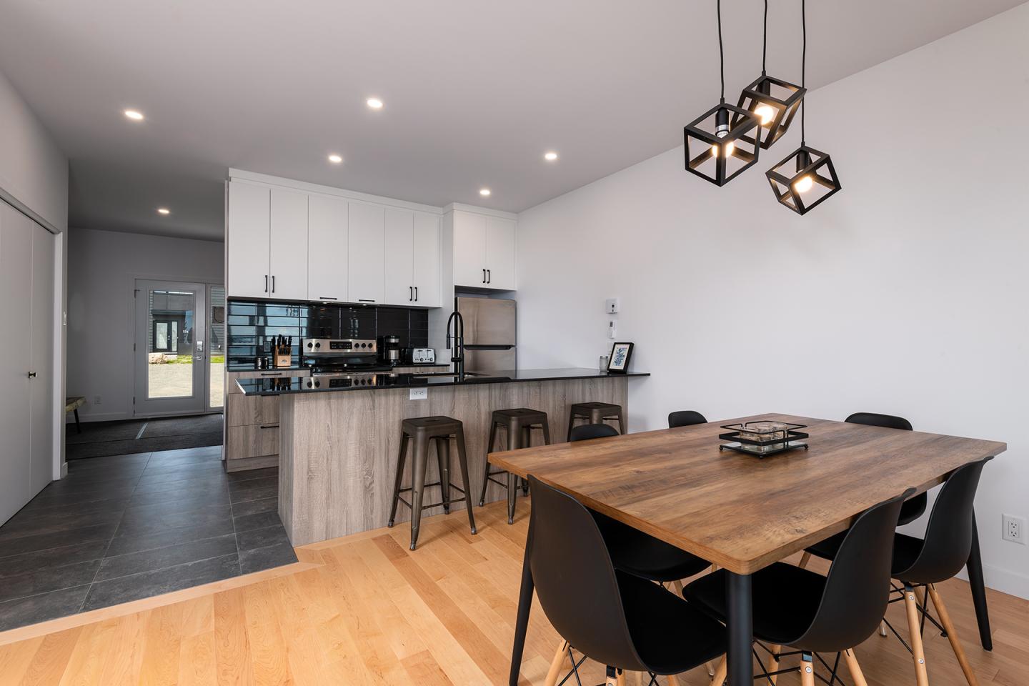 Cuisine moderne avec îlot, table en bois et chaises noires. Lumières suspendues.