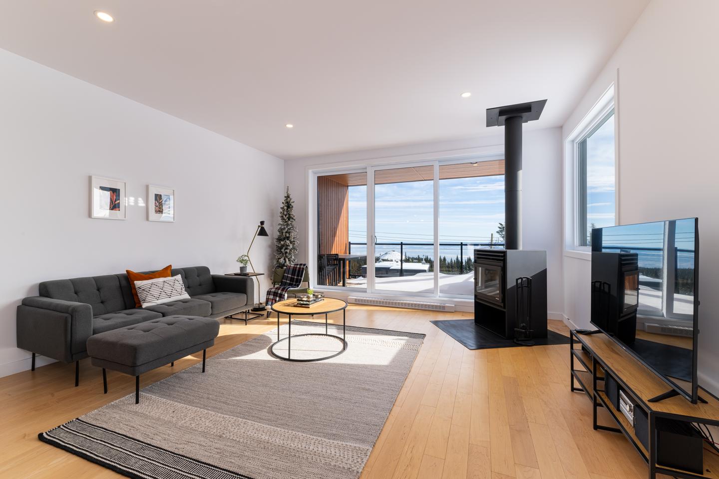 Salon moderne avec canapé gris, cheminée, grandes fenêtres et vue sur mer.