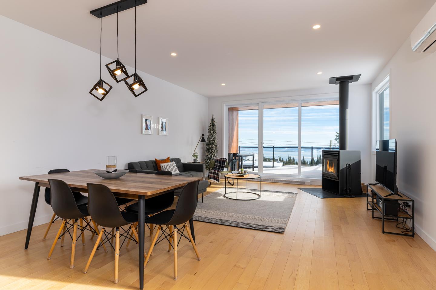 Salle à manger moderne avec table en bois, canapé, poêle et vue sur la mer.