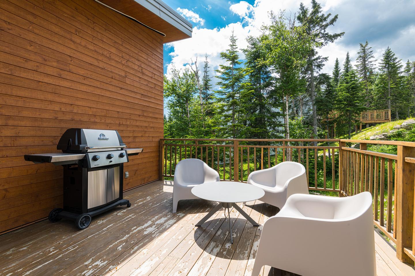 Terrasse en bois avec barbecue, deux chaises blanches et table ronde, forêt en arrière-plan.