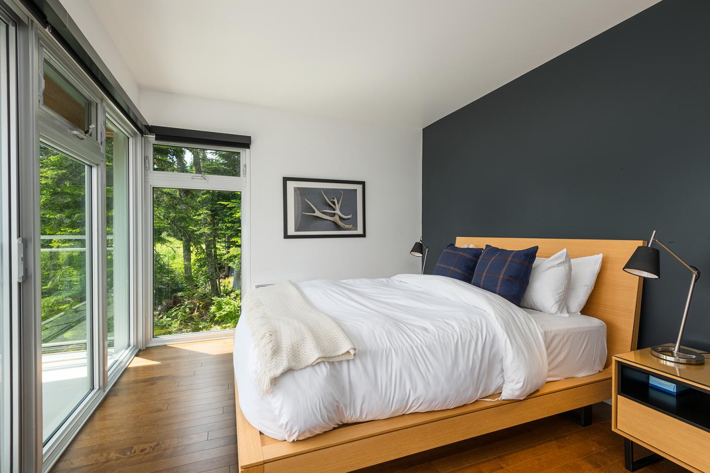 Chambre moderne avec lit double, murs sombres, grandes fenêtres et vue sur la forêt.