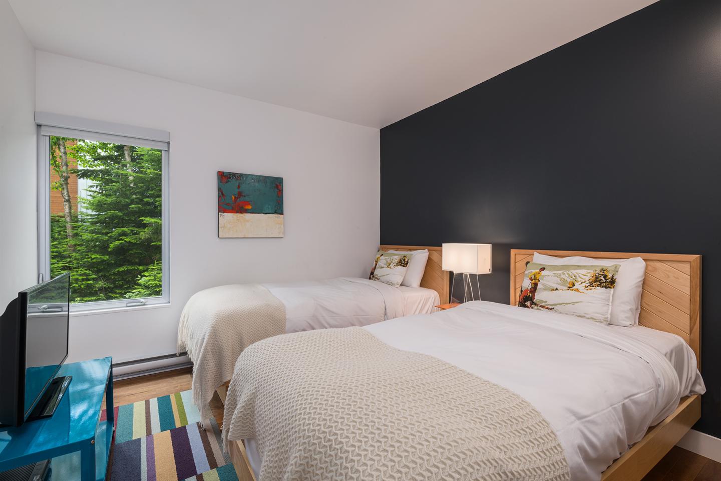Chambre moderne avec deux lits, mur bleu, et fenêtre avec vue sur des arbres.