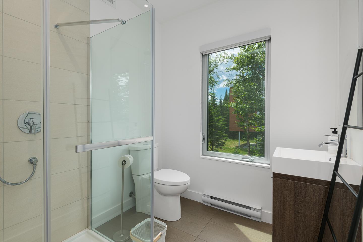 Salle de bain lumineuse avec douche vitrée, toilettes, lavabo moderne et fenêtre donnant sur la verdure.