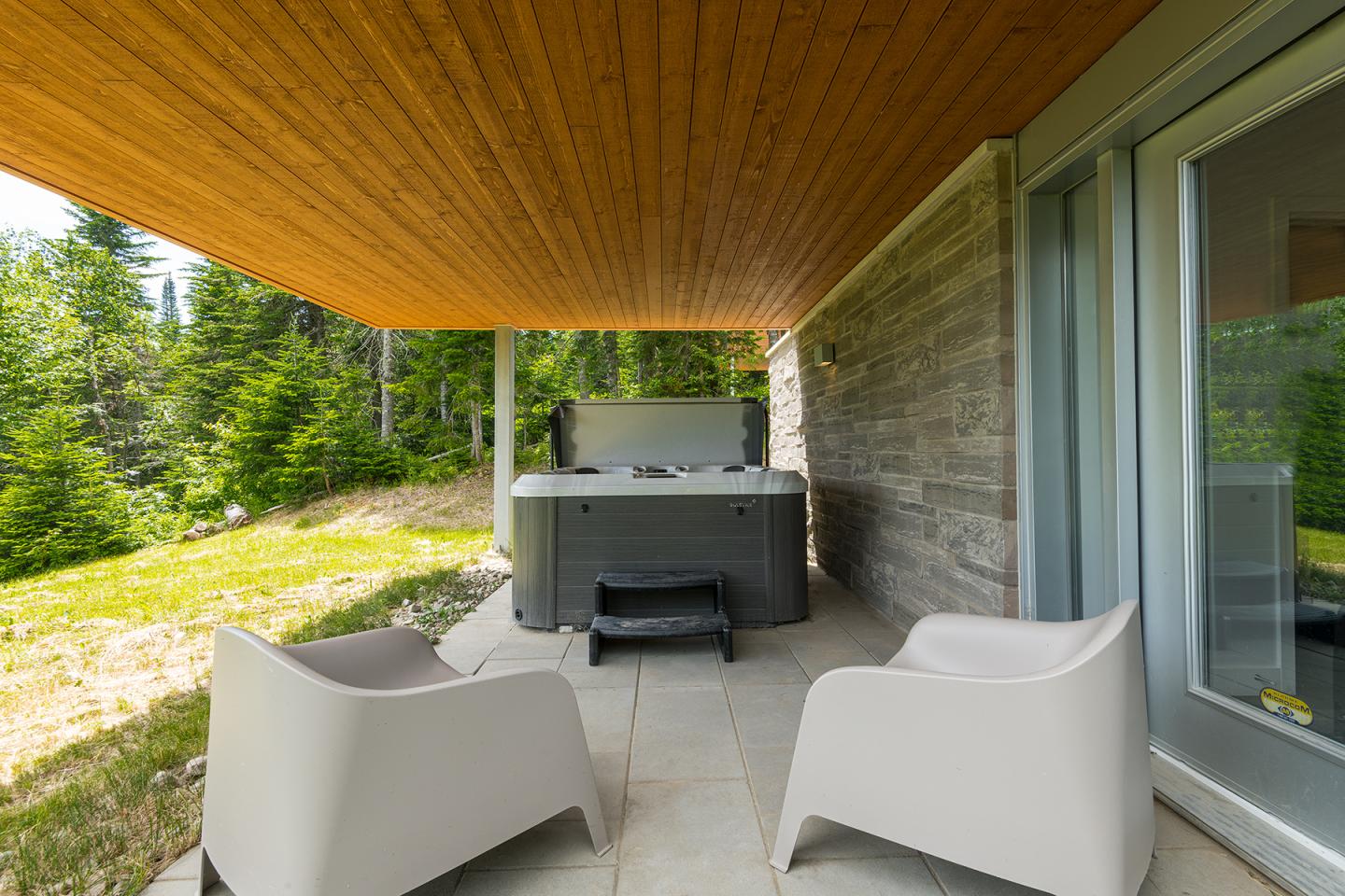 Terrasse couverte avec spa et deux chaises blanches, entourée de verdure.