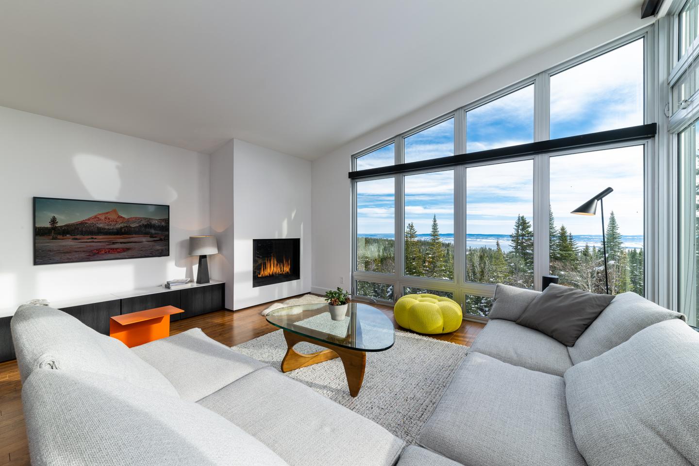 Salon moderne avec baie vitrée, canapé blanc, cheminée et vue sur la nature.