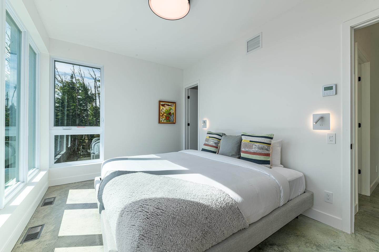 Chambre lumineuse avec grand lit, fenêtres sur la gauche, murs blancs et petit tableau coloré.