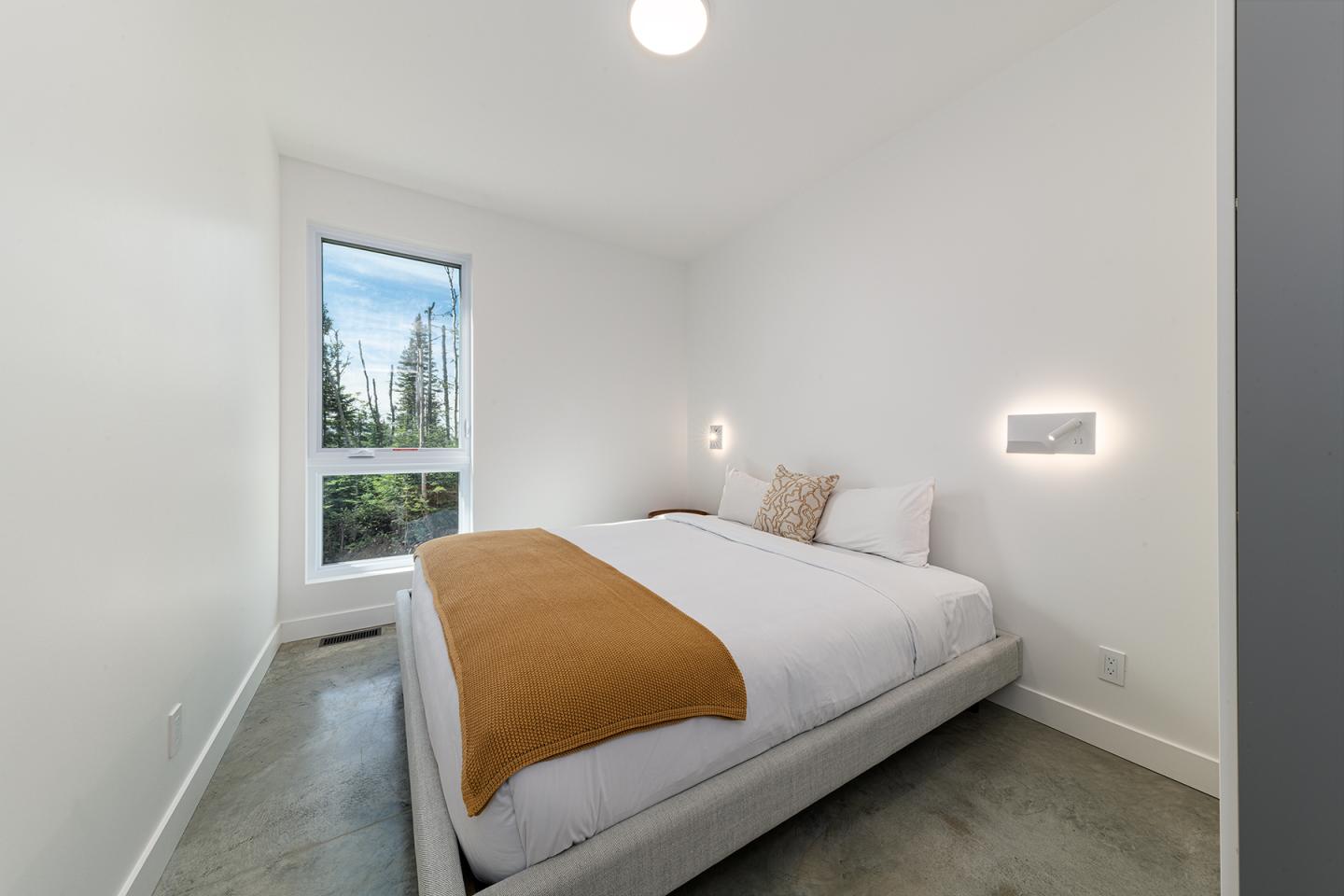 Chambre minimaliste avec lit double, couverture orange et fenêtre avec vue extérieure.