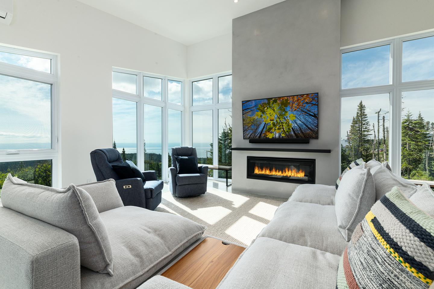 Salon moderne avec grandes fenêtres, cheminée et vue sur la forêt.