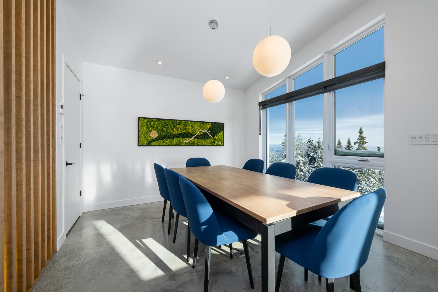 Salle à manger moderne avec table en bois et chaises bleues, grandes fenêtres avec vue.