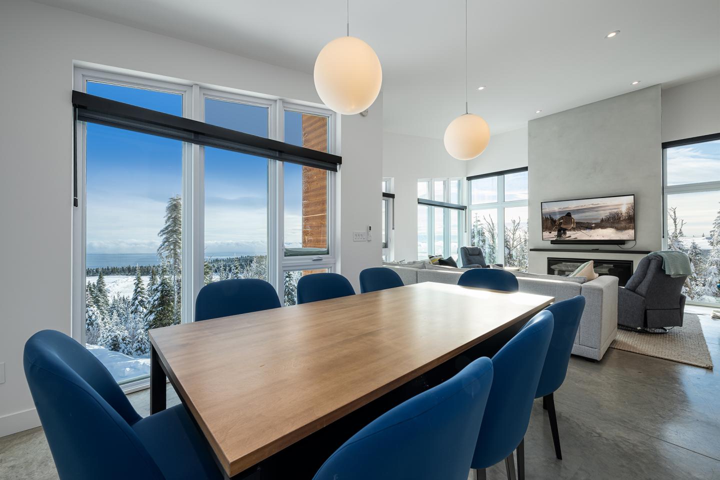 Salle à manger moderne avec table en bois, chaises bleues, baies vitrées et vue sur la neige.