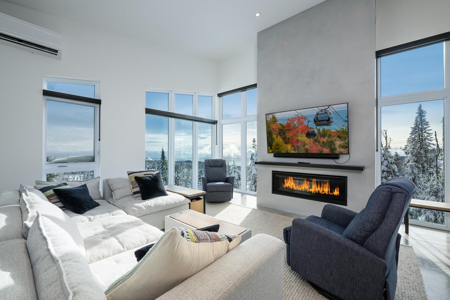 Salon moderne avec canapé blanc, cheminée, fauteuils sombres et grandes fenêtres.