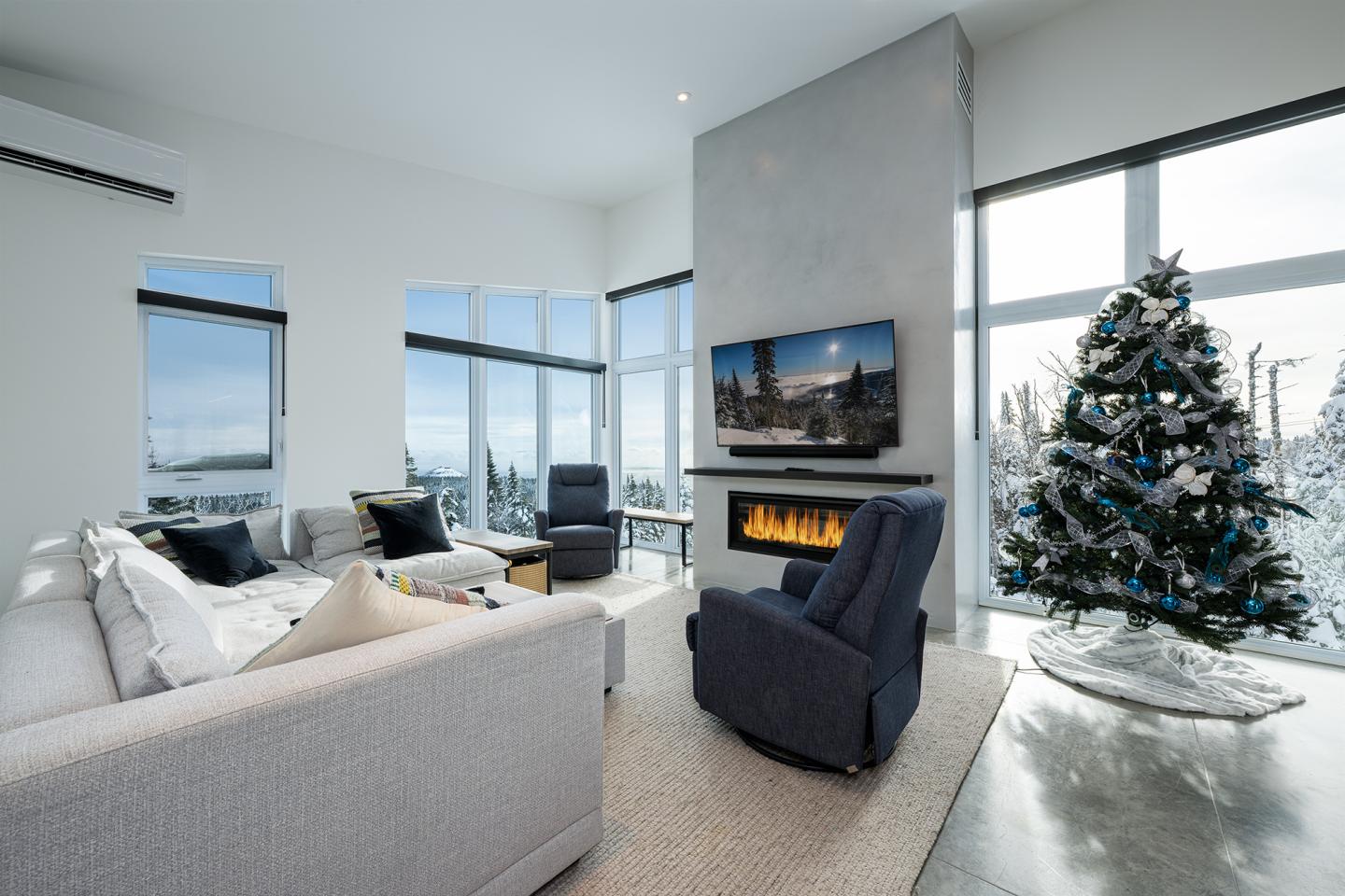 Salon moderne avec sapin de Noël, cheminée allumée et grandes fenêtres.