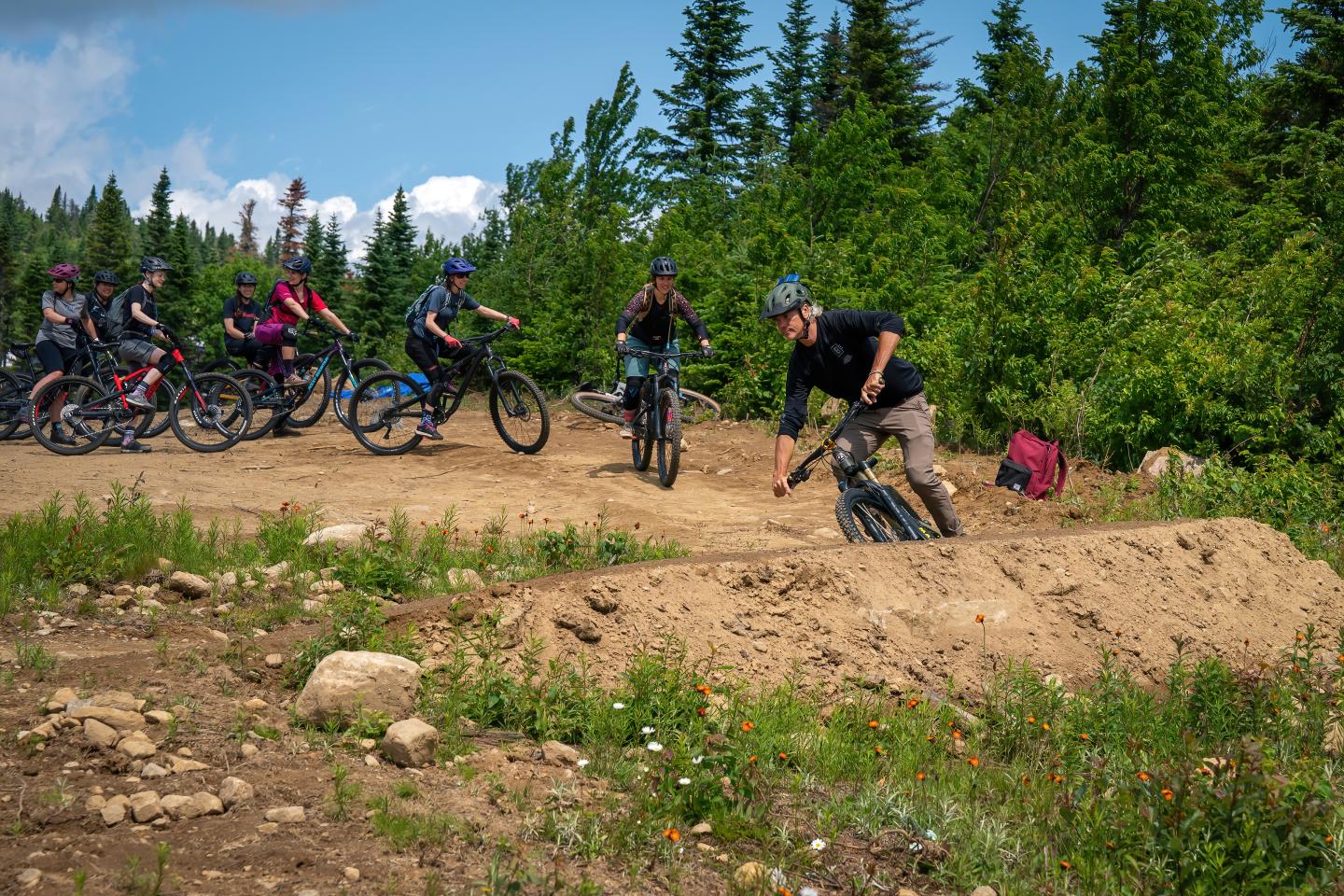 Cycliste sautant sur un sentier de terre, groupe observant à l'arrière-plan, forêt verdoyante.