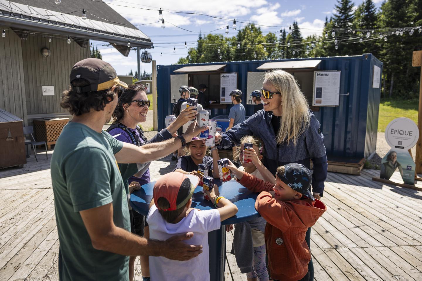 Famille et amis trinquent sur une terrasse ensoleillée, entourés de nature verdoyante.
